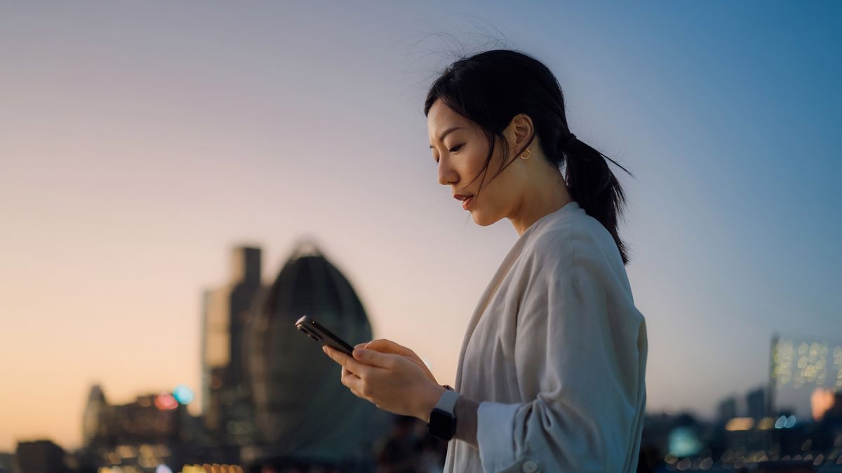 A young woman looking at her phone with an urban cityscape in the background at sunset