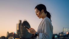 A young woman looking at her phone with an urban cityscape in the background at sunset