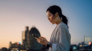 A young woman looking at her phone with an urban cityscape in the background at sunset