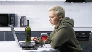 A woman is using laptop and drinking wine , in the kitchen.