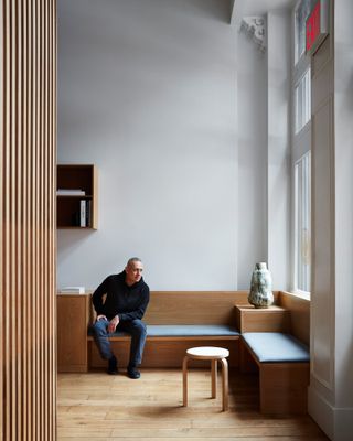 Architect Kazem Naderi sitting on a light blue upholstered bench in front of a grey wall, in the wooden space he created for Spiral (x,y,z)