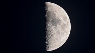 The moon half-lit moon is pictured against a dark sky at its first quarter phase. Dark lunar seas mark the upper portion of the lunar surface and the line dividing the night side from the day side is lined with shadowed craters.