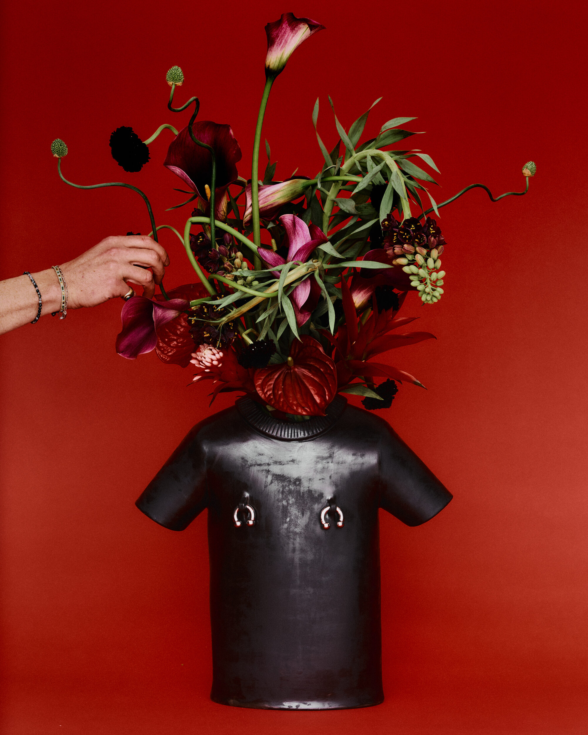 Image of a flower vase that is shaped like a black t-shirt in front of a red background. There are green, red, and pink flowers in the vase and a hand reaching towards it.