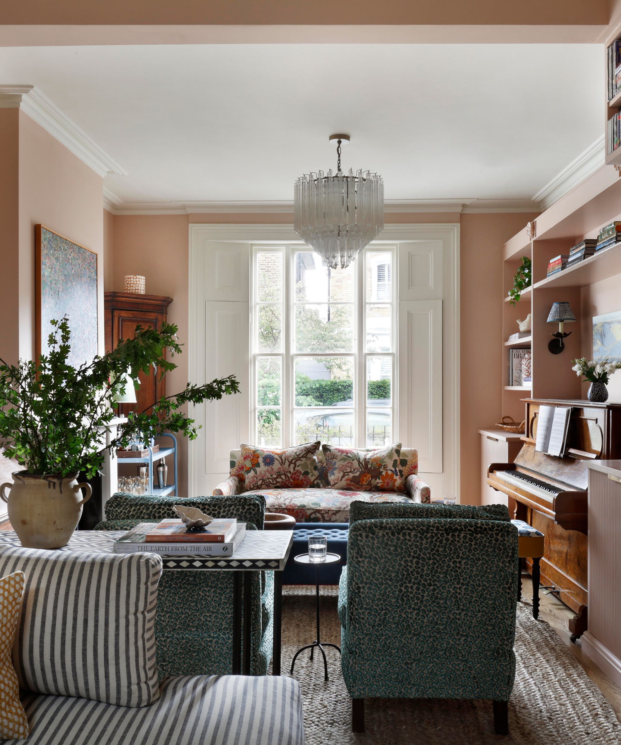 Warm, blush-pink living room with a crystal chandelier, a upright piano, floral and patterned armchairs, and a large bay window flanked by white shutters.