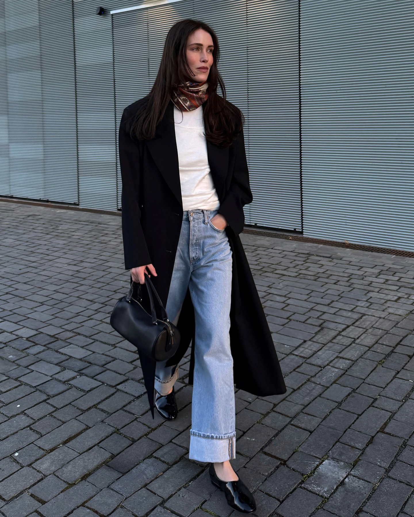 style influencer Annabel Rosendahl walking wearing a white T-shirt, black lightweight coat, silk printed neck scarf, black bag, cuffed jeans, and black glove flats