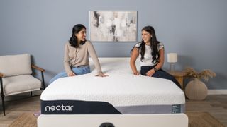 Two girls sitting on Nectar Premier memory foam mattress in sleep studio with blue wall