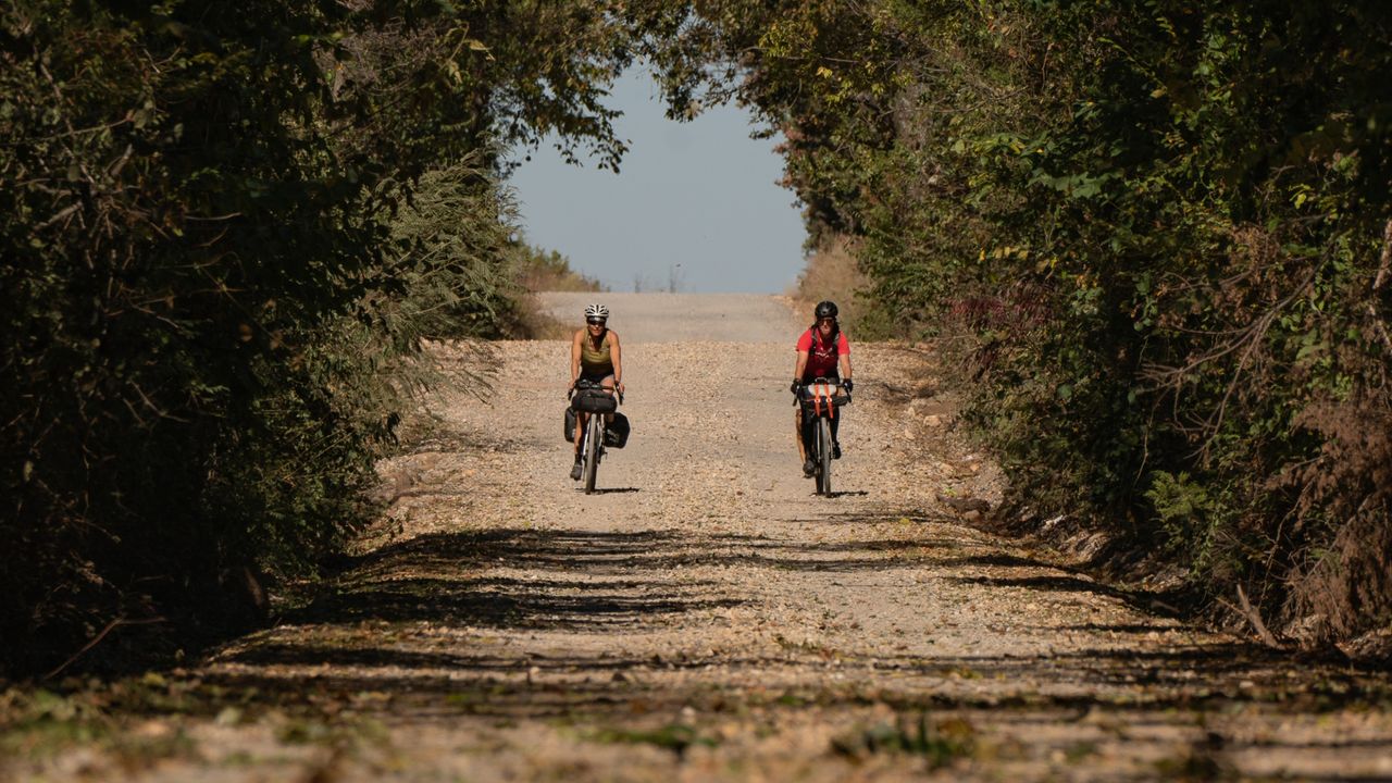 A man and woman ride gravel bikes down a tan gravel road in Arkansas, towards the camera.