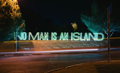 Outdoor night time image, quiet road, grass lawns, green neon floor standing letters, spelling 'NO MAN IS AN ISLAND' , trees, dark sky, silhouette trees & landscape in the distance
