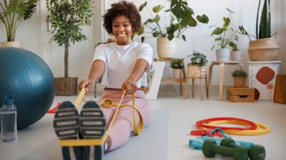 A woman sits with her legs straight in front of her and a resistance band looped round her feet; she is holding the ends of the resistance band taught in her hands. She smiles straight ahead. Beside her, we see an exercise ball, more resistance bands and a set of light dumbbells. Behind her we see lots of potted plants.