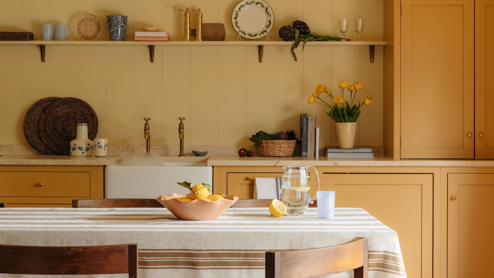 butter yellow kitchen with soft green panelled walls, deeper yellow shaker cabinets, farmhouse sink, open shelving and a kitchen table styled with a linen and brown striped tablecloth