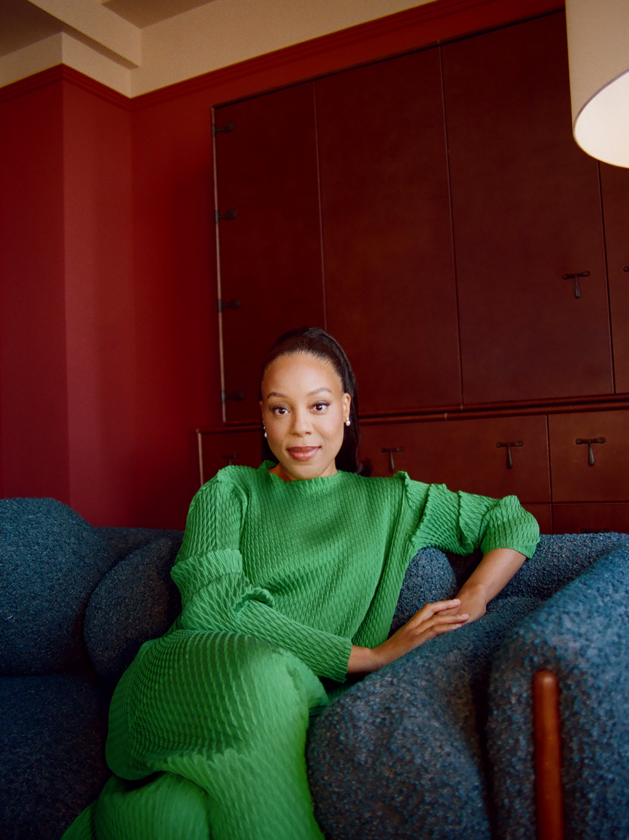 Orion Pictures president Alana Mayo wears a green sweater dress while sitting in a blue felt chair in front of a hotel room's wooden armoir.
