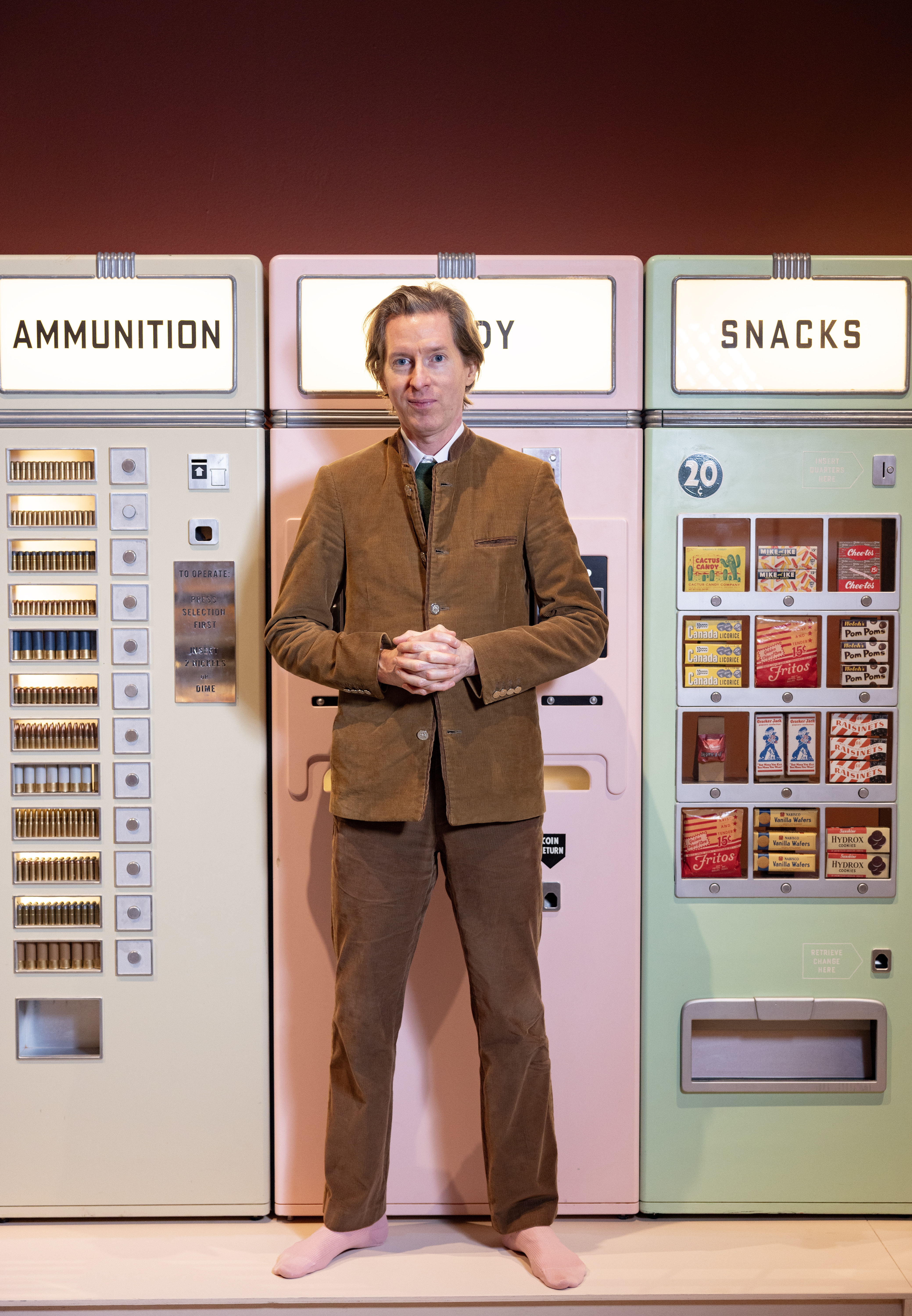 A middle-aged man dressed in a non-matching, velvety brown suit stands with his hands crossed in front of his chest in front of a series of pastel-shaded food vendors with a vintage look, painted beige, pink, and mint green.