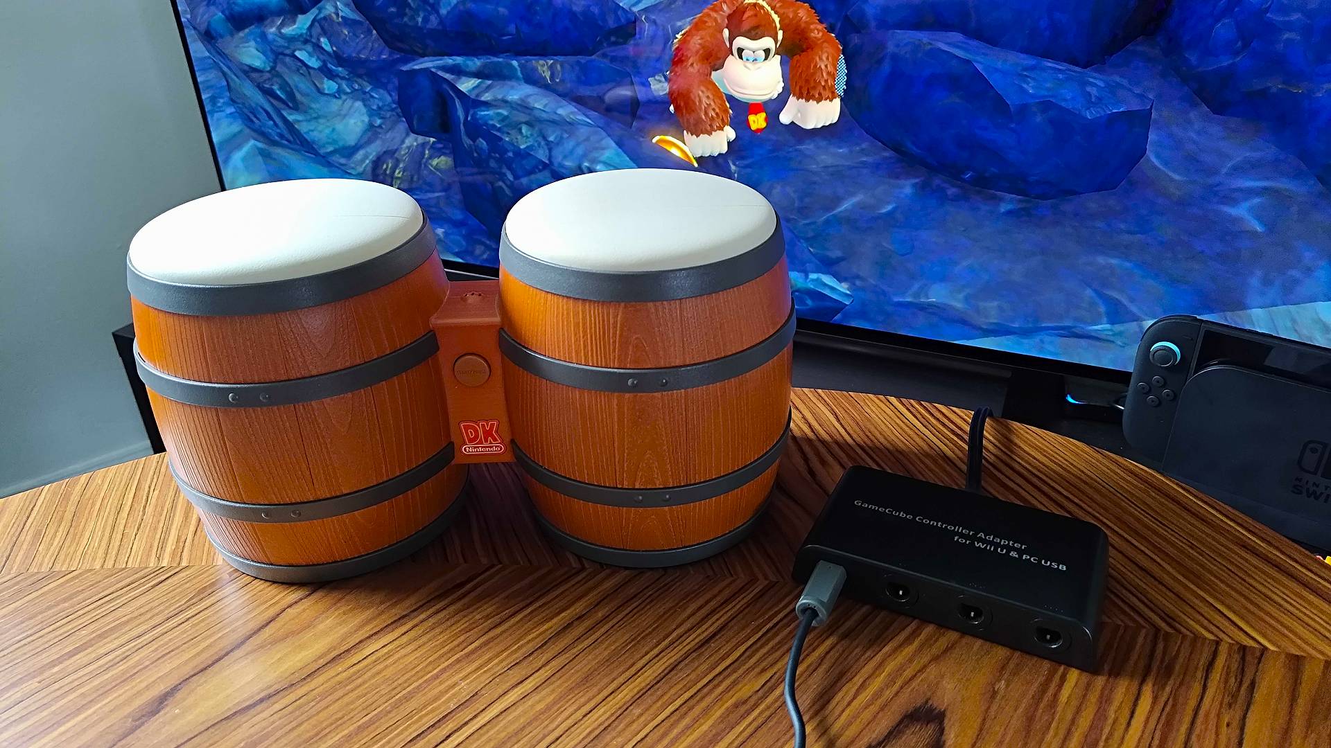 Donkey Konga Bongos next to Mayflash Gamecube adapter on table with Donkey Kong Bananza gameplay in backdrop on TV.