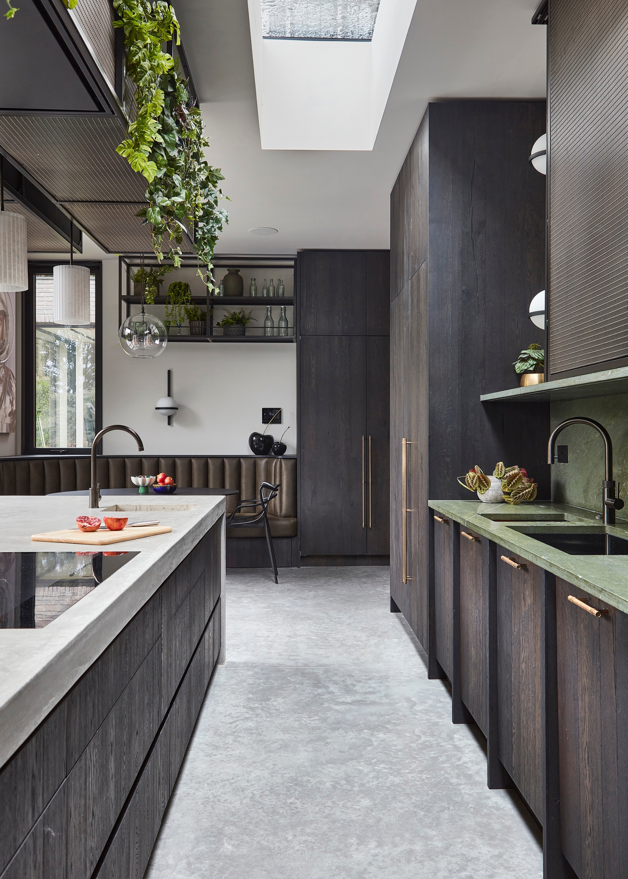 A dark wood off-black kitchen with brass hardware, green stone worktops, brown banquette seating, and a concrete look floor