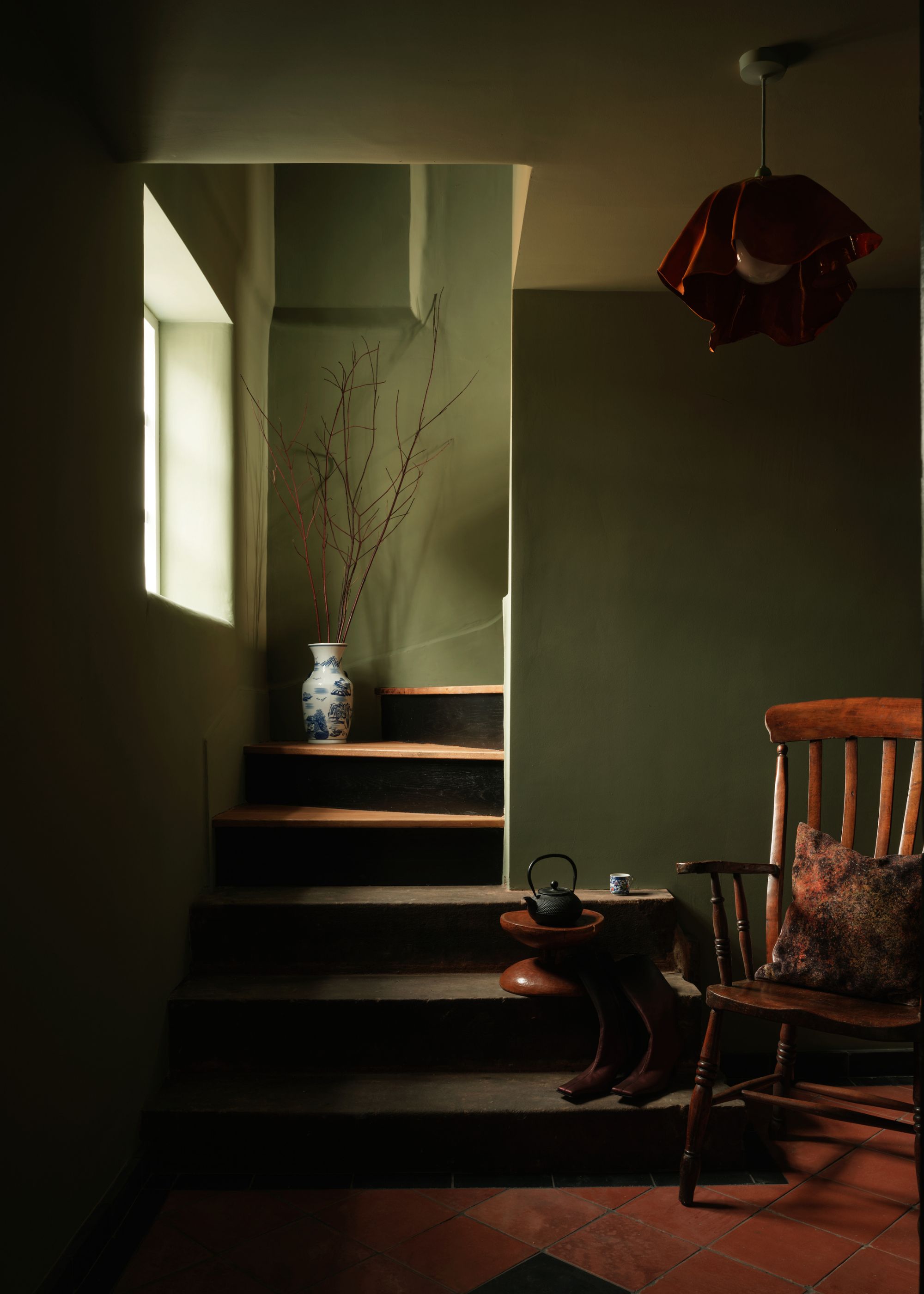 A green color drenched area of the home with a staircase leading to another area of the home. In the middle of the staircase is a vase with branches inside it. In this room there is also a wooden chair with a cushion on top as well as a mini side table and shoes sitting on one of the staircases.