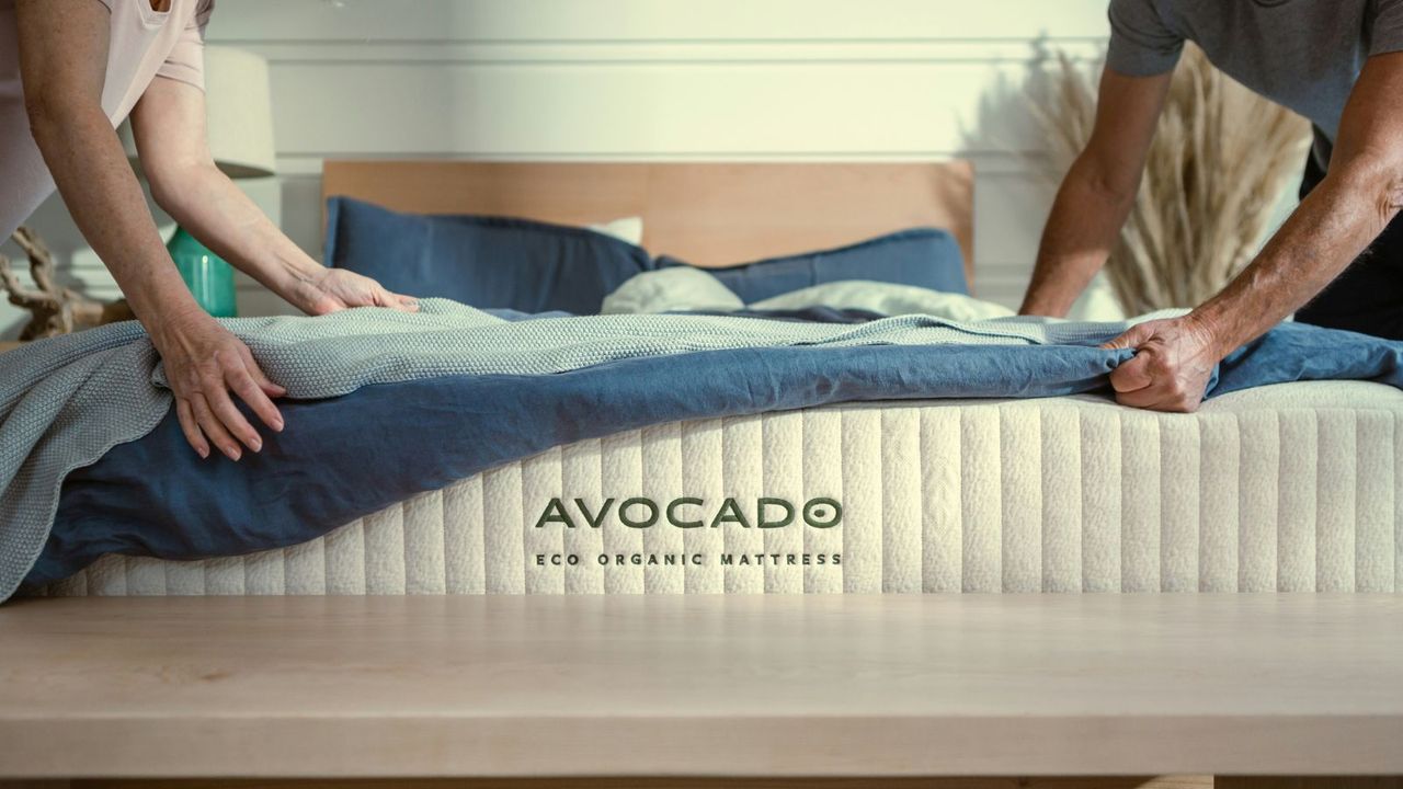 Hands pulling a blue duvet and light blue blanket over the Avocado Eco Organic Mattress. 