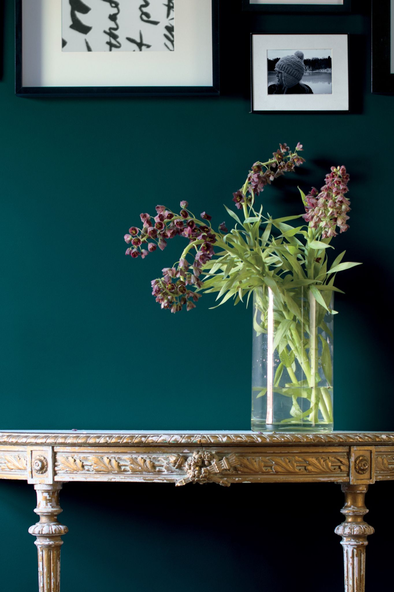 Jade green painted wall with a gallery wall of black and white pictures and a bronze antique table with a floral arrangement