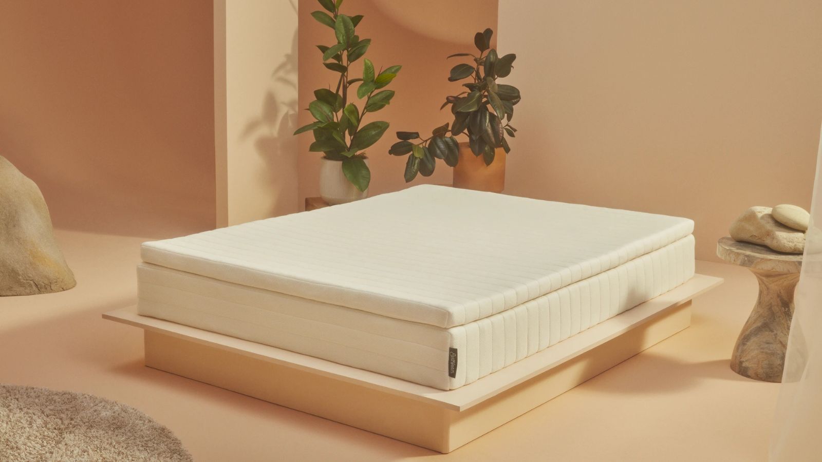 Double bed with exposed mattress and matter topper, beige painted walls and floor, plants, marble side table