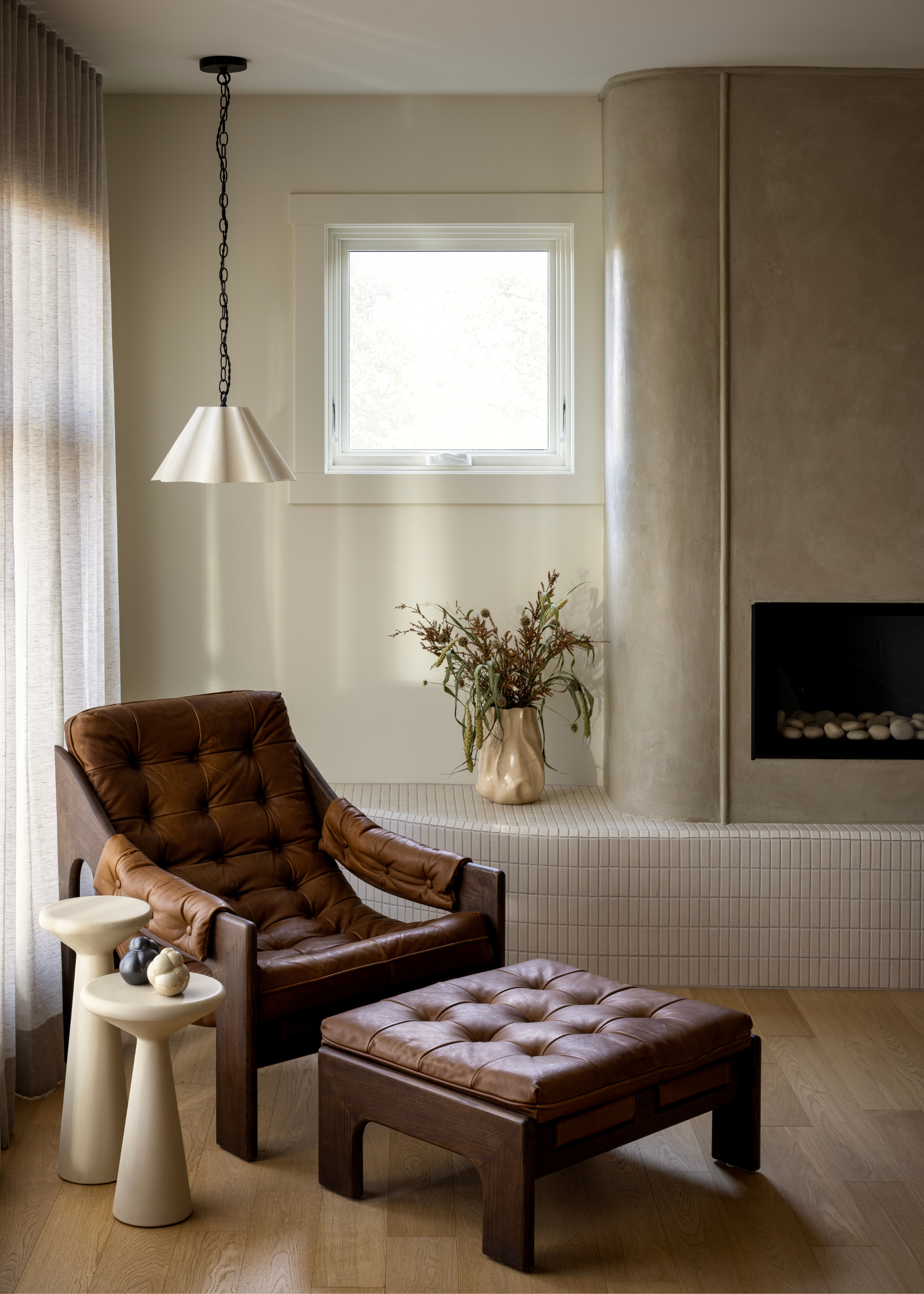 A living room corner with a leather chair and foot stool, by a pair of white side tables, a tiled extended fireplace, a vase of dried flowers and a hanging pendant