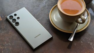 En ljusgrå Samsung Galaxy S23 Ultra ligger på ett mörkt bord bredvid en tekopp med baksidan vänd uppåt.