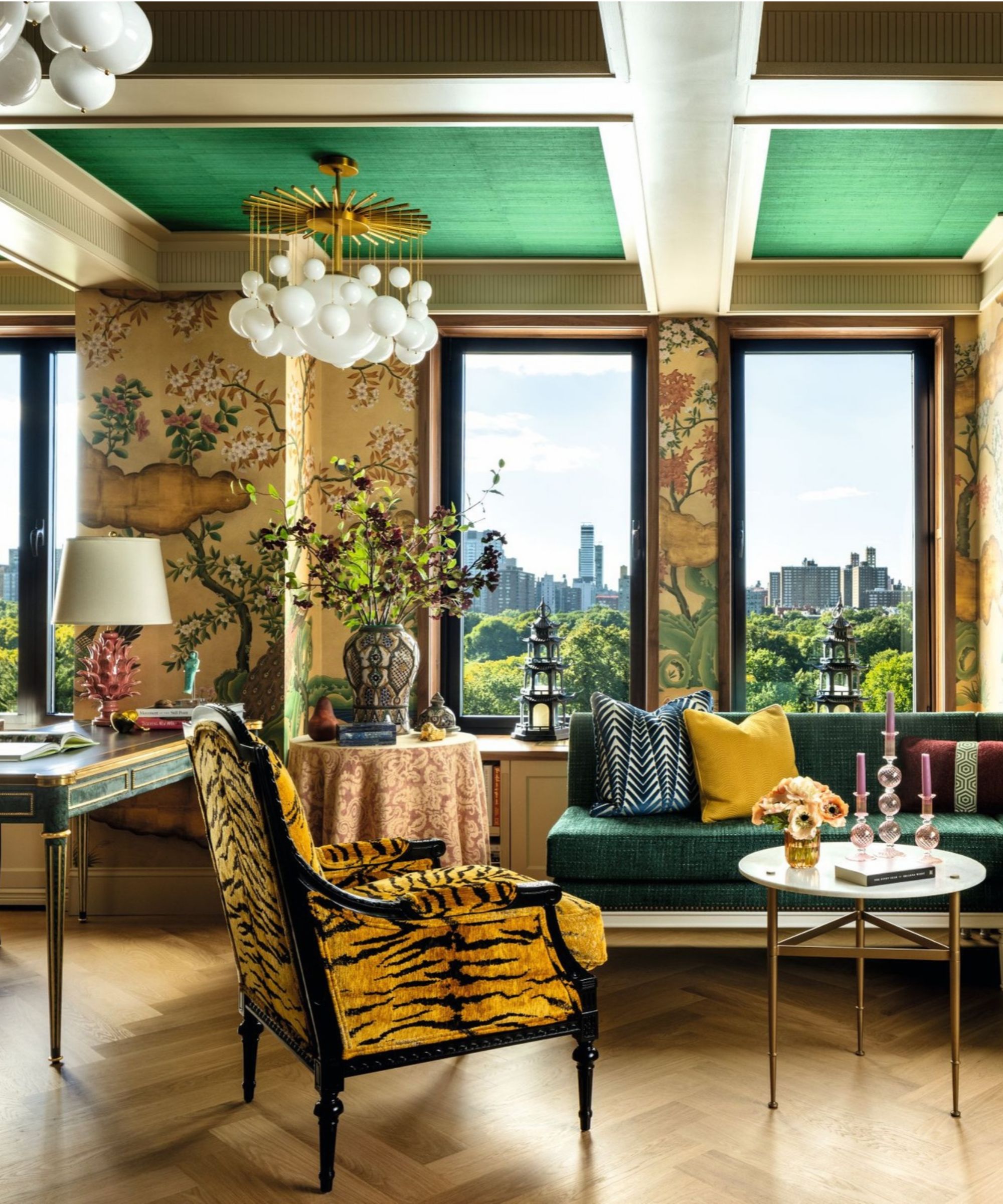 Living room with tiger print chair in foreground, forest scene wallpaper, a desk to left with lamp, an central round table with a vase of flowers, a green sofa to the right with a small side table