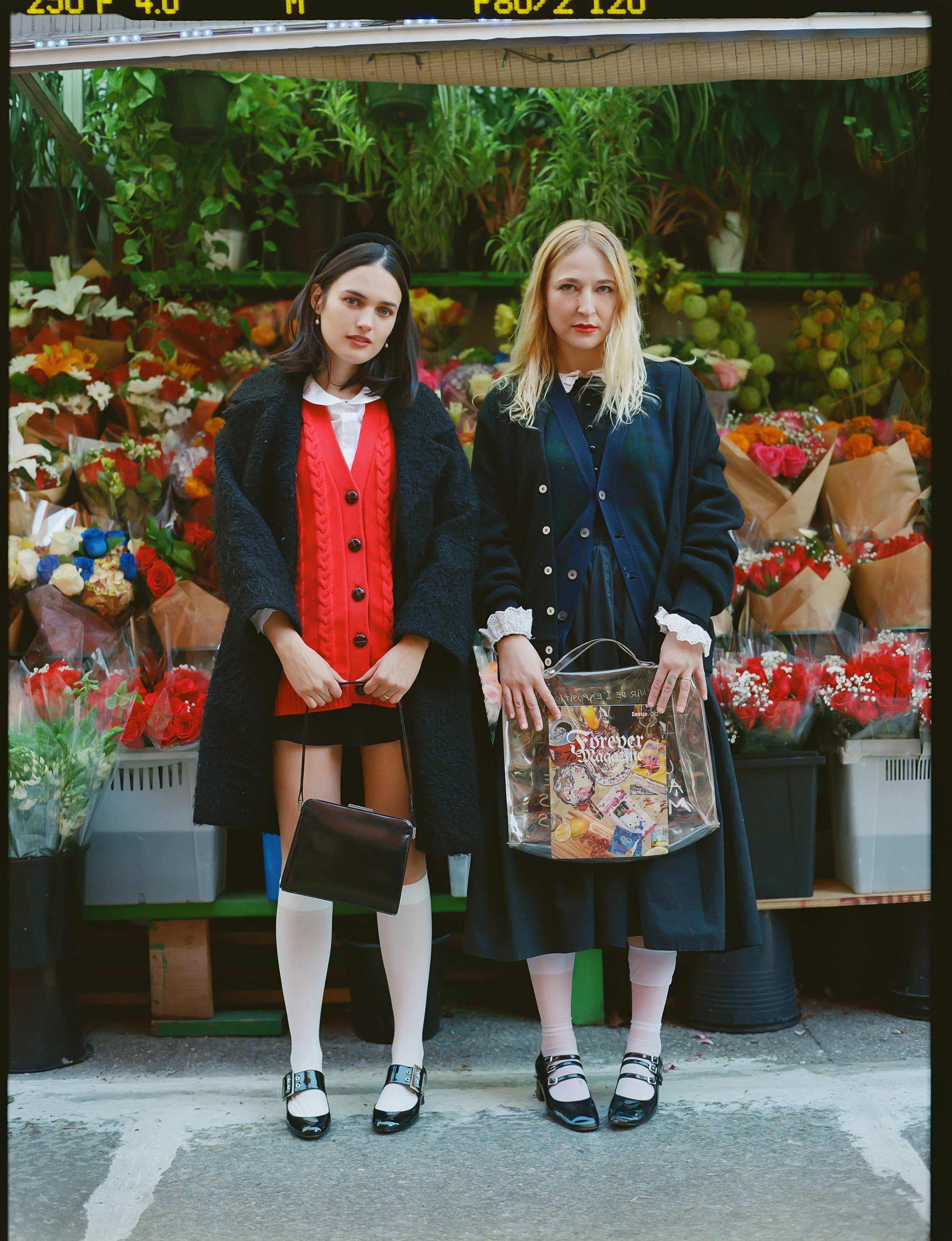 forever magazine cofouners nat ruiz and anika jade levy pose in front of bodega flowers wearing preppy outfits with white socks and mary janes and hold a copy of their mag