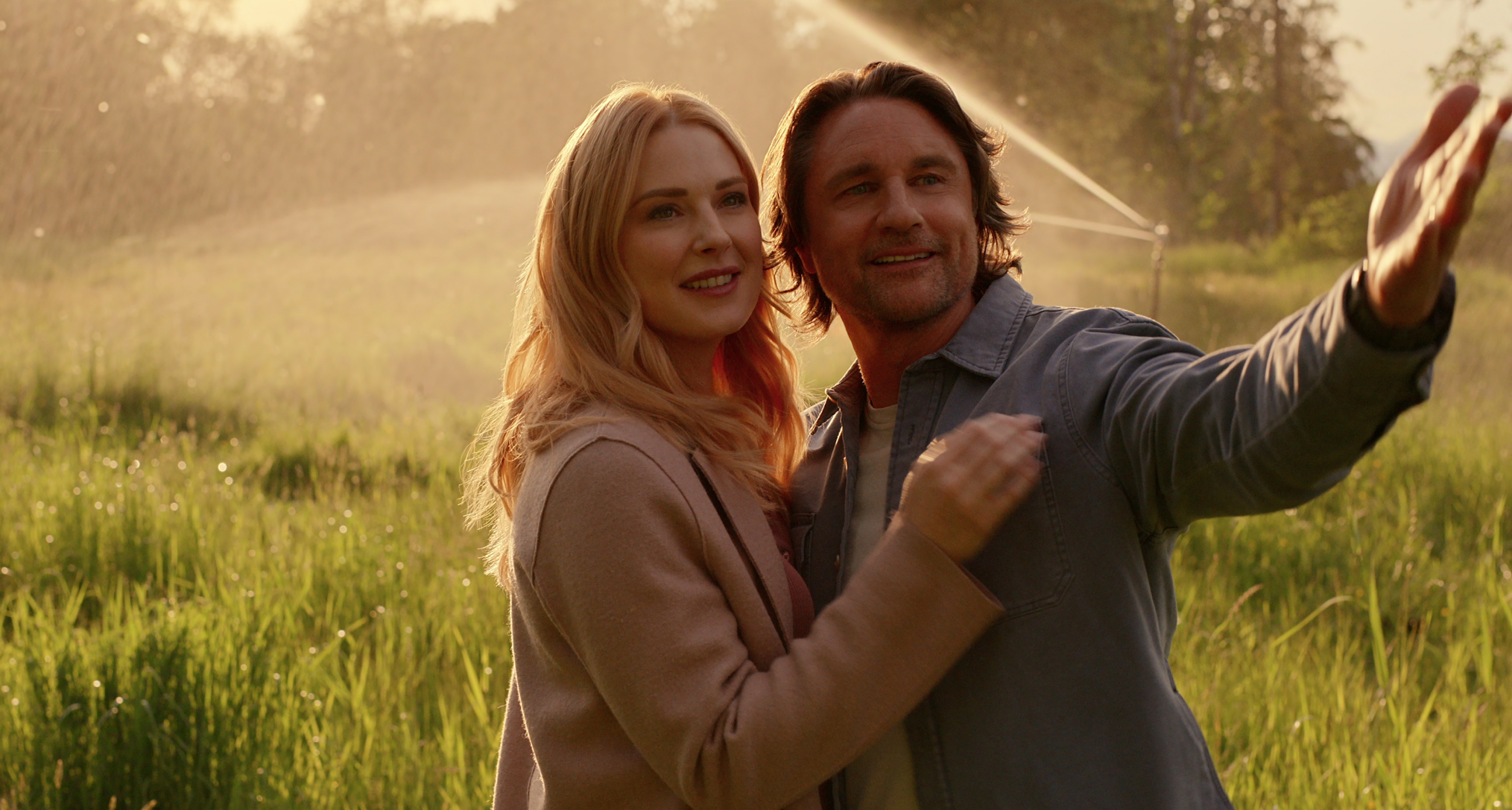 alexandra breckenridge as melida and martin henderson as jack smiling and looking out into a field while a sprinkler goes off behind them in a still from virgin river season 7