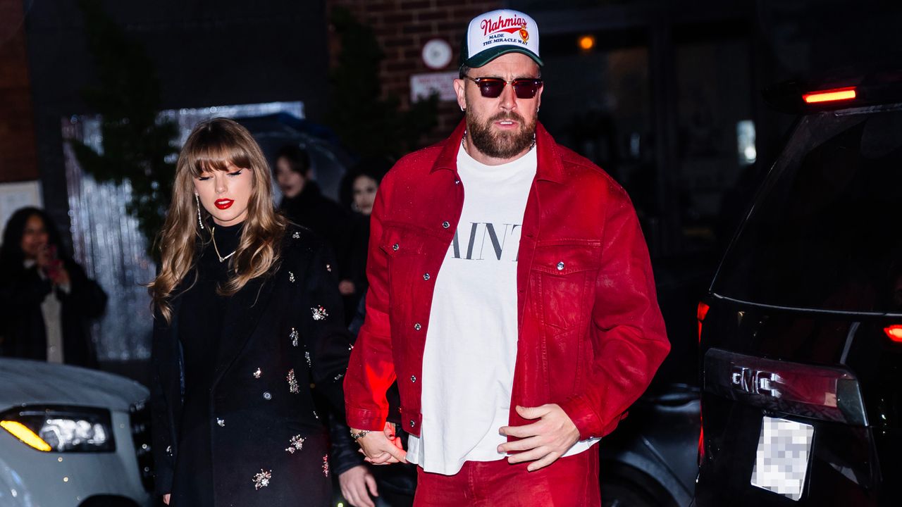 Travis Kelce wearing a red jacket and white tee walking next to Taylor Swift in a black coat walking into a restaurant