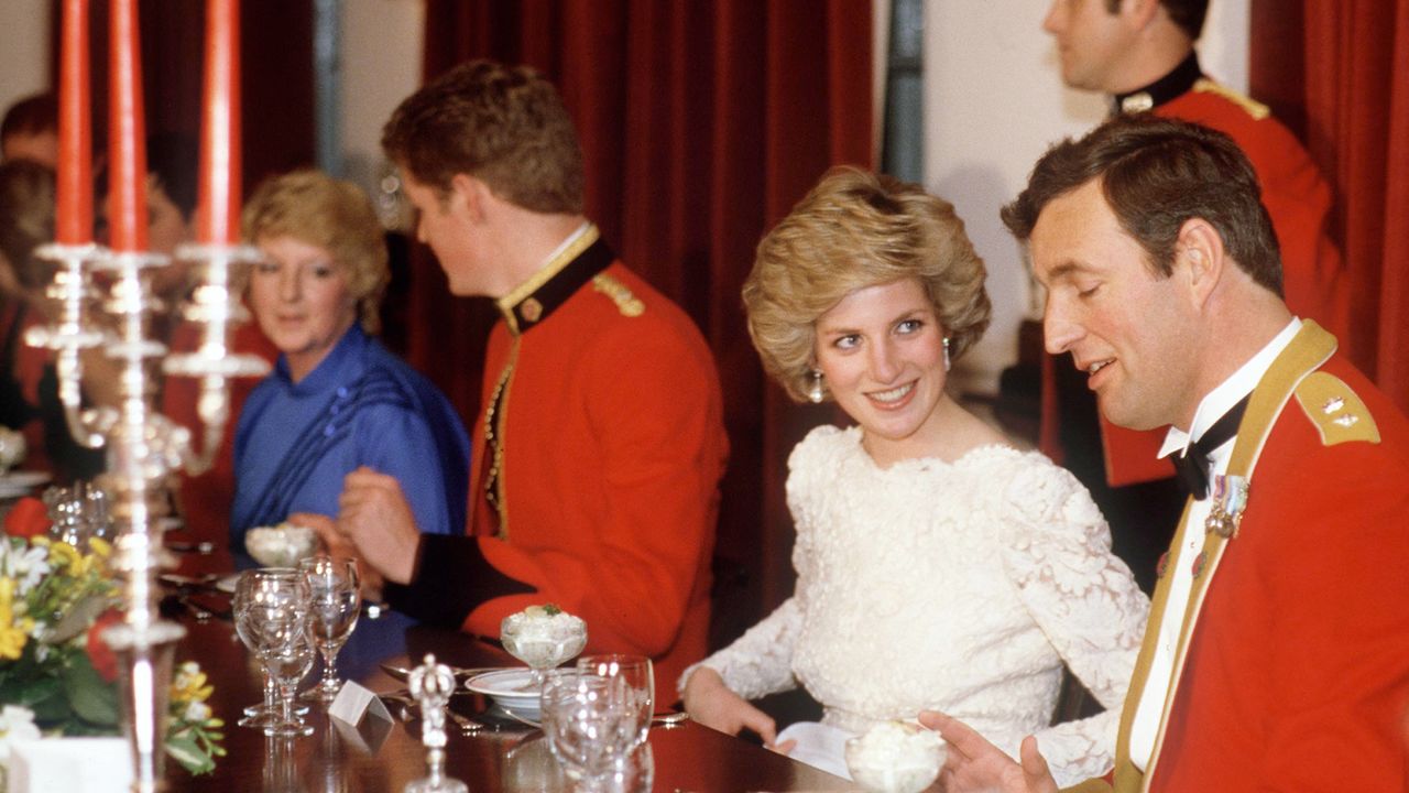 Princess Diana wearing a white lace gown sitting at a banquet table between two soldiers in red uniforms