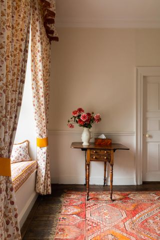 stone bedroom with window seat and upsholstered cushion fall drapes, orange, red and yellow vintage rug, antique side table with vase of dahlias