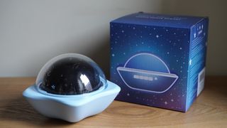 A dome-shaped projector on a white star-shaped stand, in front of a blue box, on a brown wooden surface.