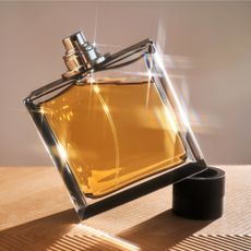 Image of a sunlit perfume bottle filled with gold liquid, stood at an angle on the lid and set against a wood and taupe wall background