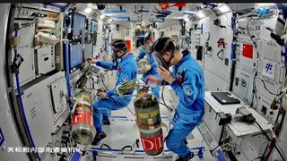 Three astronauts in blue jumpsuits float in a white space module, wearing gas masks connected to gas tanks.