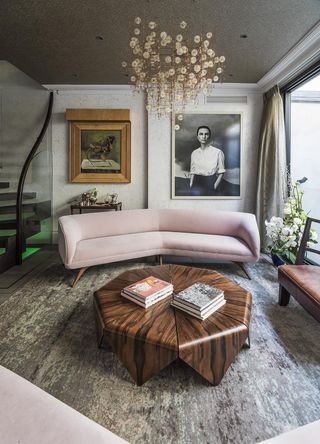A lounge area with a pink sofa, a leather chair, an octagonal wooden coffee table, potted plants, wall paintings and a chandelier.