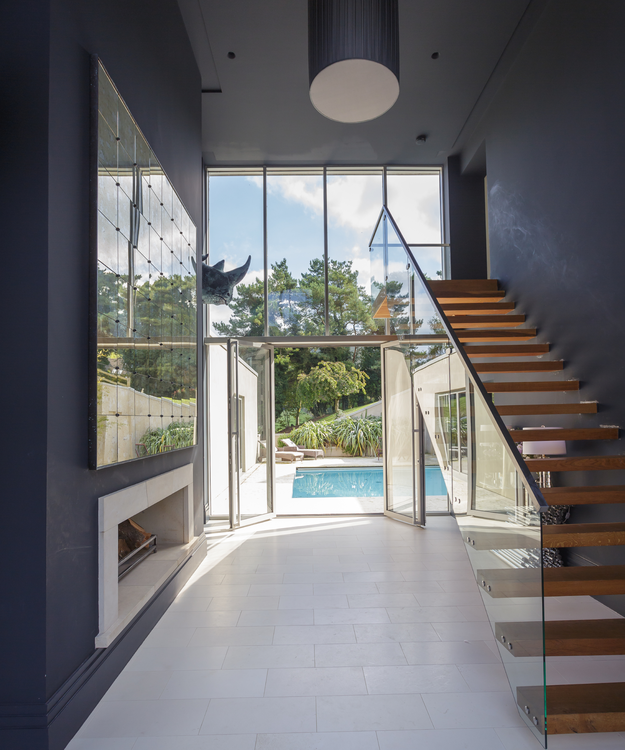inside of modern home with double height hallway featuring floating wooden stairs with glass balustrade, large patio doors at end leading to outside swimming pool