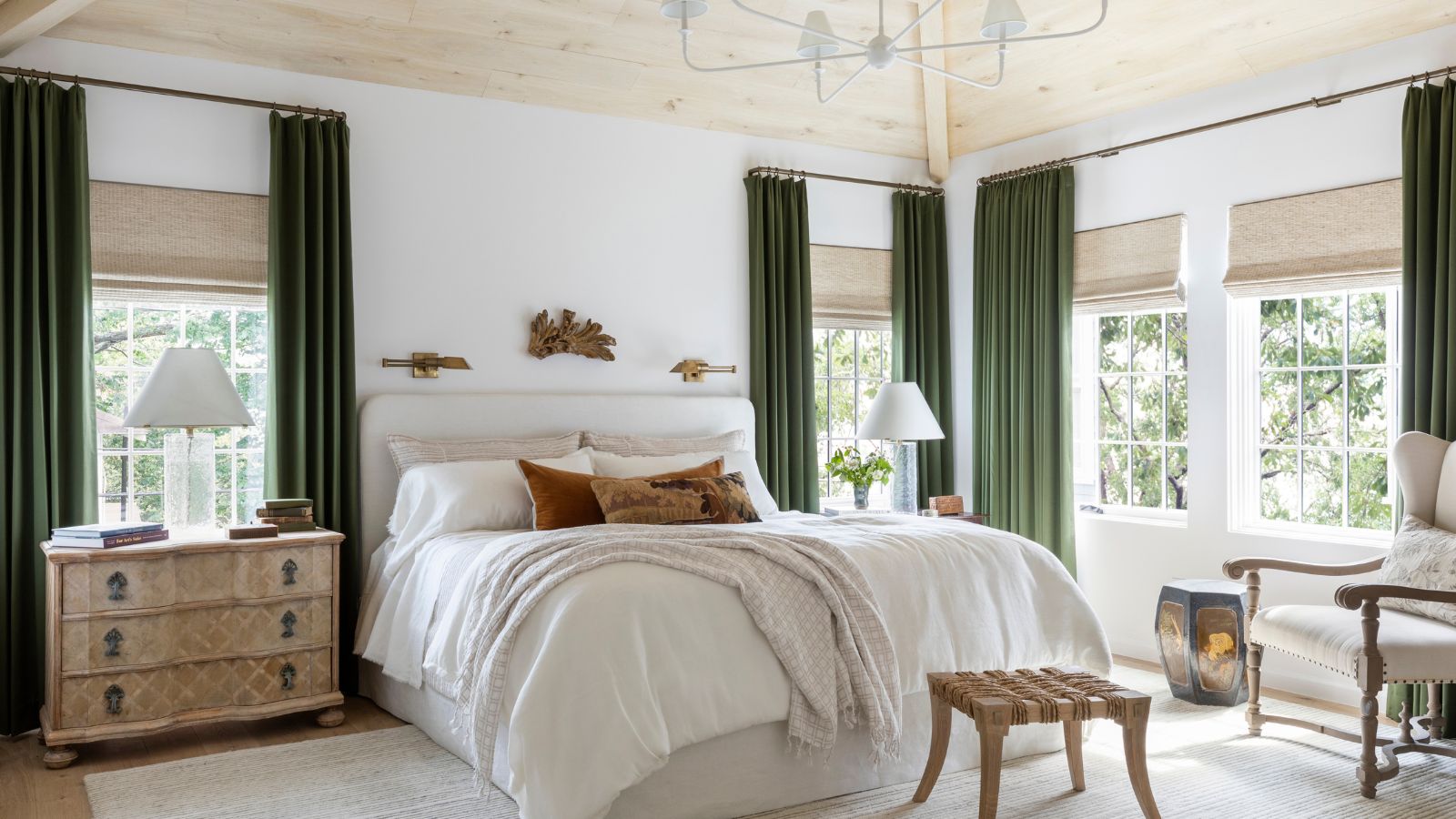 A modern light and airy bedroom with a pale wood-clad ceiling, white walls, a large beige rug, olive green curtains, and a large bed with layered neutral bedding. 