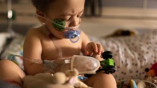 photo of a toddler recovering from rsv in a hospital bed; he's sitting up and holding a toy care while sucking on a binky. An oxygen tube is inserted into his nostrils and secured to his face with colorful adhesive bandages