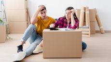 Young couple sitting amid moving boxes with a calculator, figuring out the cost of moving.