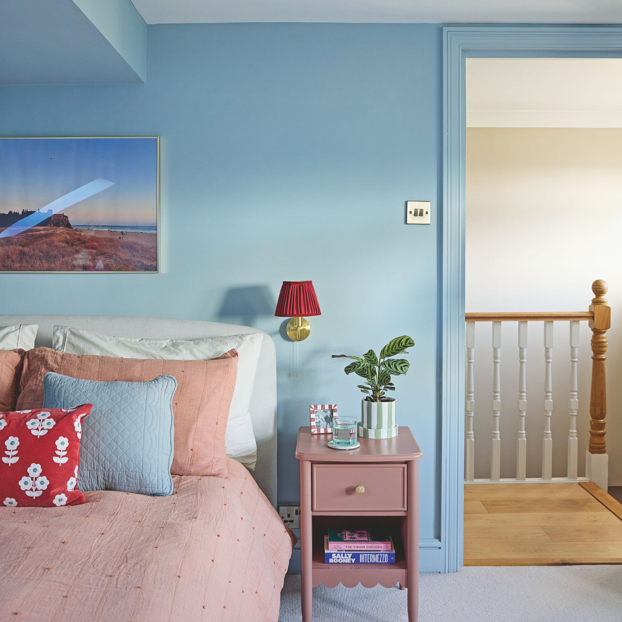 Blue painted bedroom off a landing area with wooden floors. There is pink bedding on the bed and a pink side table next to it