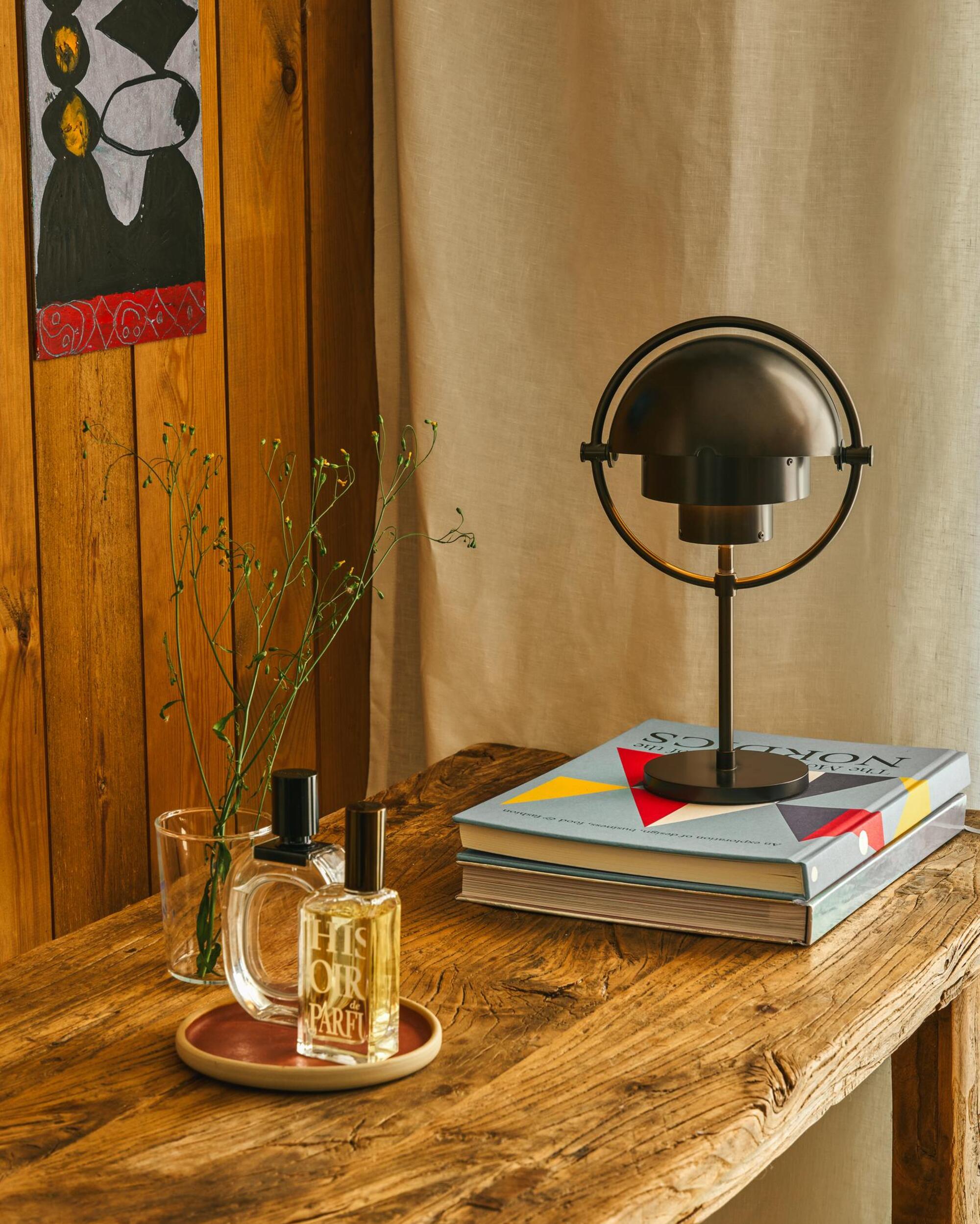 gubi rechargeable lamp sitting on timber desk and stack of books, next to a dish holding perfumes and a vase of flowers