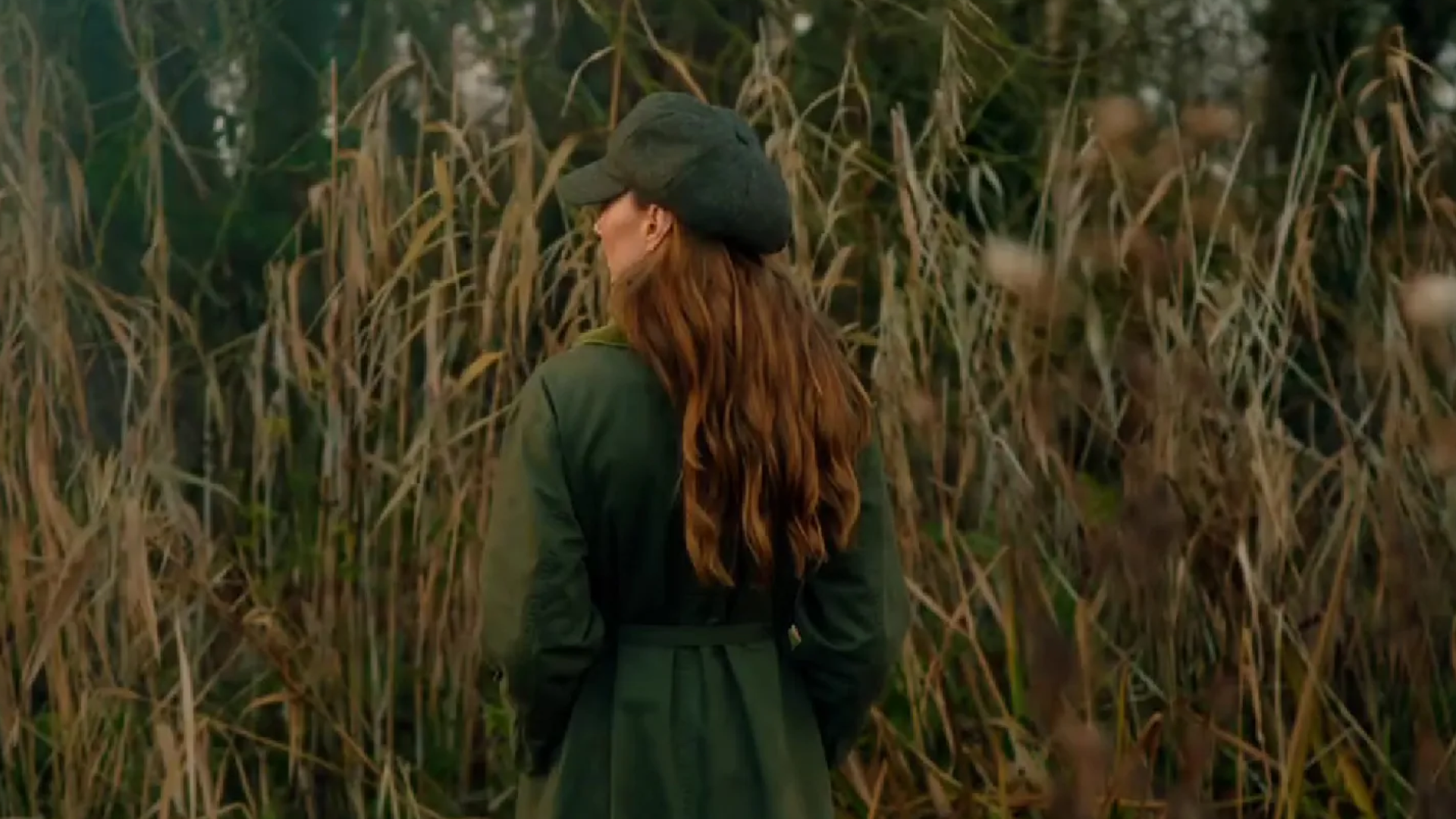 Kate Middleton standing in front of tall grass in a green coat and hat with her back to the camera