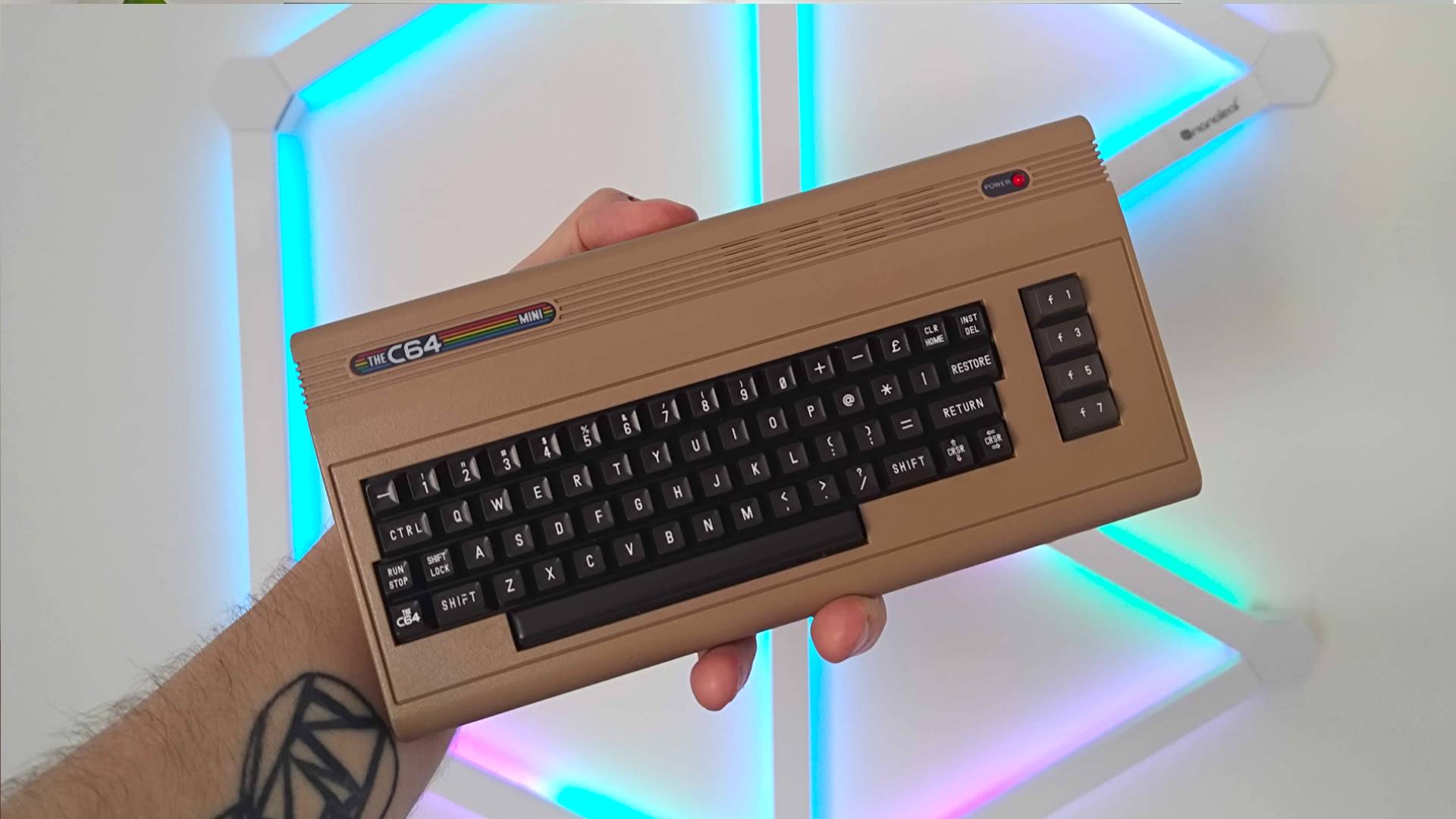 Hand holding The C64 Mini console with RGB blue and pink lights in backdrop.