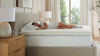 A woman laying on Saatva graphite mattress topper on thick white mattress in light bedrrom with big windows looking out to greenery, wooden bedside table and lamps