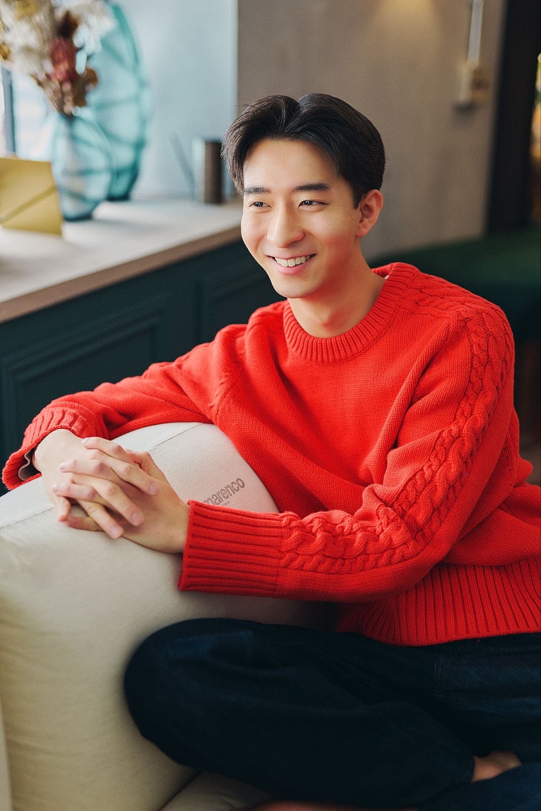 A Japanese man named Bomi wears a red sweated and black pants while sitting on a couch with his hands laced on a pillow, in his cast portrait for 'The Boyfriend.'