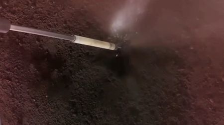 a long, thin metal tube extends into a black-grey dusty surface, and a cloud of dust and sparks forms where the two meet