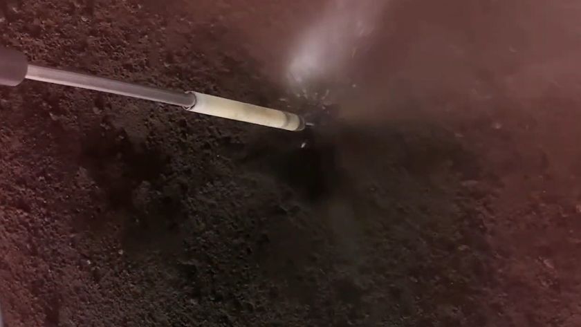 a long, thin metal tube extends into a black-grey dusty surface, and a cloud of dust and sparks forms where the two meet