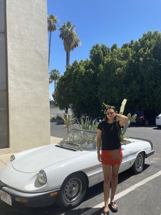 Woman in an orange skirt, flip-flops, and black shirt standing in front of a car with cacti.