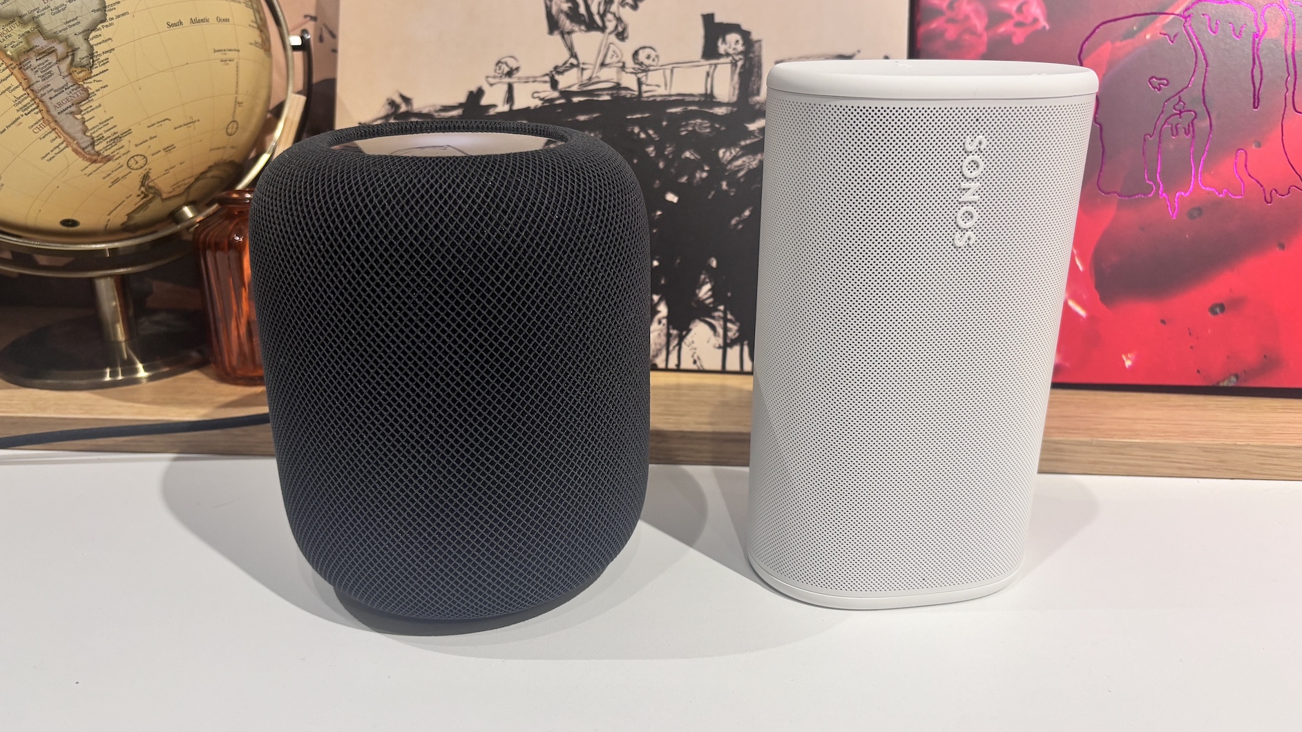 A black Apple HomePod 2 standing next to a white Sonos Play smart speaker on a white surface in front of vinyl sleeves.
