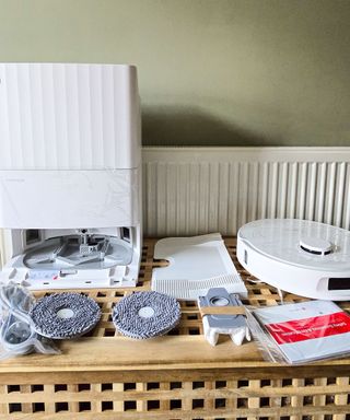 A wooden table with a a white robot vacuum base, two mop heads, a robot vacuum, and instructions on top of it, with a green wall behind it