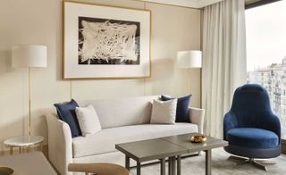 Interior view of a living area in a hotel suite at The One, Spain featuring light coloured walls, framed wall art, two white and gold floor lamps, a grey coffee table, a white and gold side table, a dark blue and grey swivel chair, a light coloured sofa with cushions and a large window with white curtains